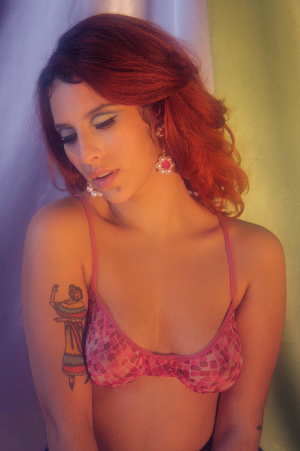 A woman with red hair and dangling pearl earrings casts her gaze towards the ground and holds a sultry pose against a purple and green backdrop. The image is softly lit giving it a slightly dream-like feeling